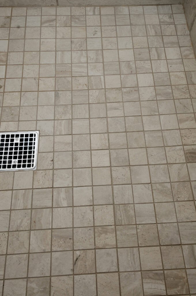 A tiled shower floor with beige square tiles and a rectangular metal drain cover on the left side.