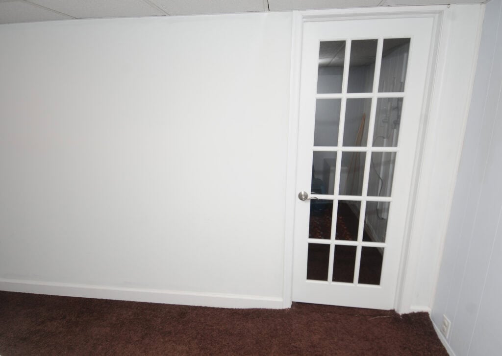 A white wall with a brown carpeted floor and a glass-paneled French door leading to another room. The area appears to be clean, empty, and well-lit.