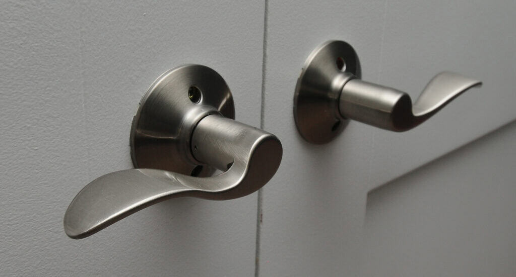 Two modern silver door handles are mounted on a gray door, positioned side by side, with smooth, curved lever designs on a flat background.