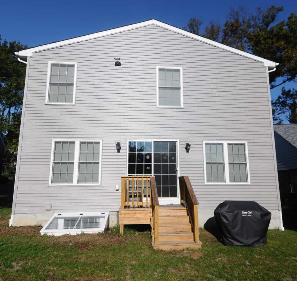 A two-story house with light gray siding, five windows, a sliding glass door, a small wooden porch with stairs, and a black grill on a grassy backyard. The sky is clear and blue.