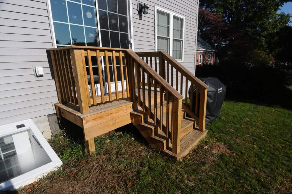 A small wooden deck with three steps and railings leads to the back door of a house. The deck sits above a grassy yard next to a grill and a basement window well.