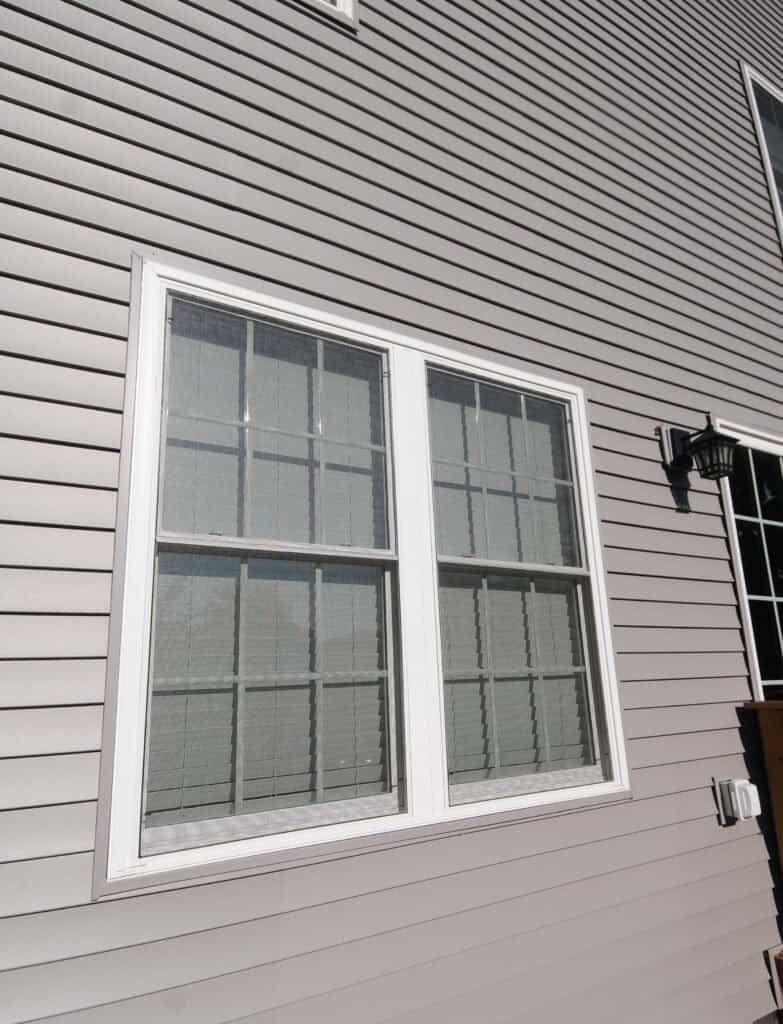 A close-up of a double window with white trim on the exterior of a gray house with horizontal siding. The window reflects sunlight and shows its grid pattern. A black outdoor light fixture is mounted nearby.