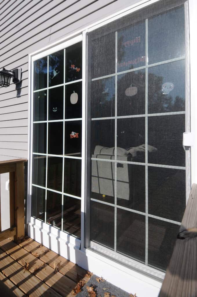 A sliding glass door with a screen door and Halloween-themed window clings, including a pumpkin and ghost, on a sunny day. A wooden deck and part of a house with gray siding are visible.