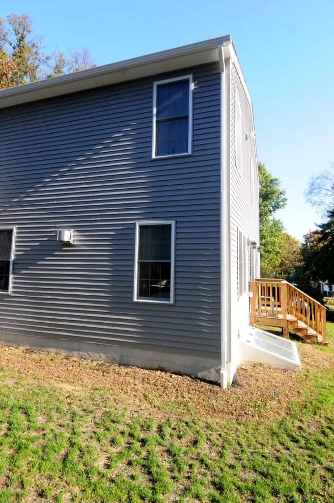A two-story house with blue-gray siding, three windows, and a small wooden ramp leading to a door. The house sits on a grassy yard with sunlight and clear blue sky.