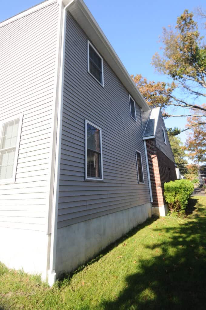 A two-story house with gray vinyl siding and brick accents, featuring several windows, sits on a grassy lawn under a clear blue sky with some trees nearby.
