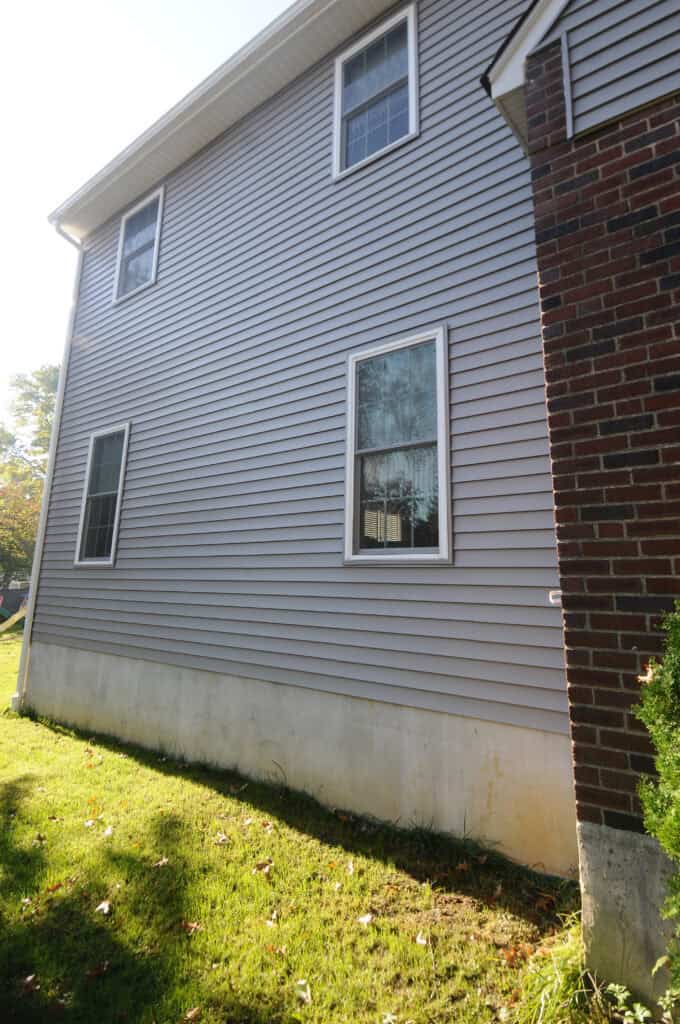 A two-story house exterior with gray vinyl siding, three windows, and a section of red brick. The lower siding shows dirt stains near the grass lawn, and sunlight casts shadows on the building.