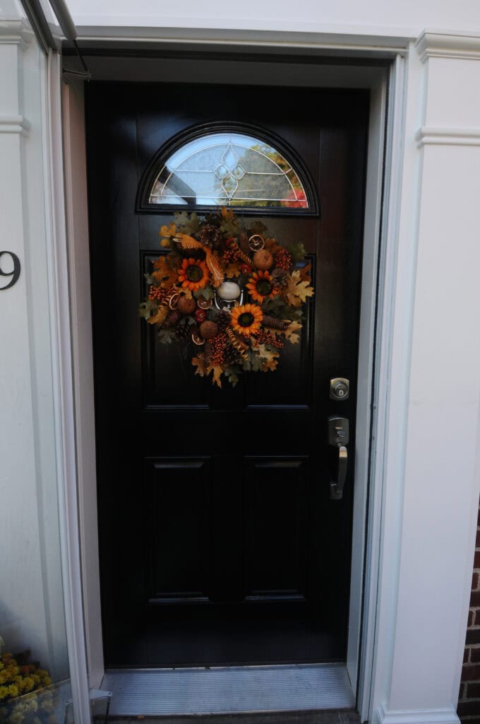 A black front door decorated with a fall-themed wreath made of orange sunflowers, leaves, and pinecones. The door has a silver handle and a semi-circular window at the top.