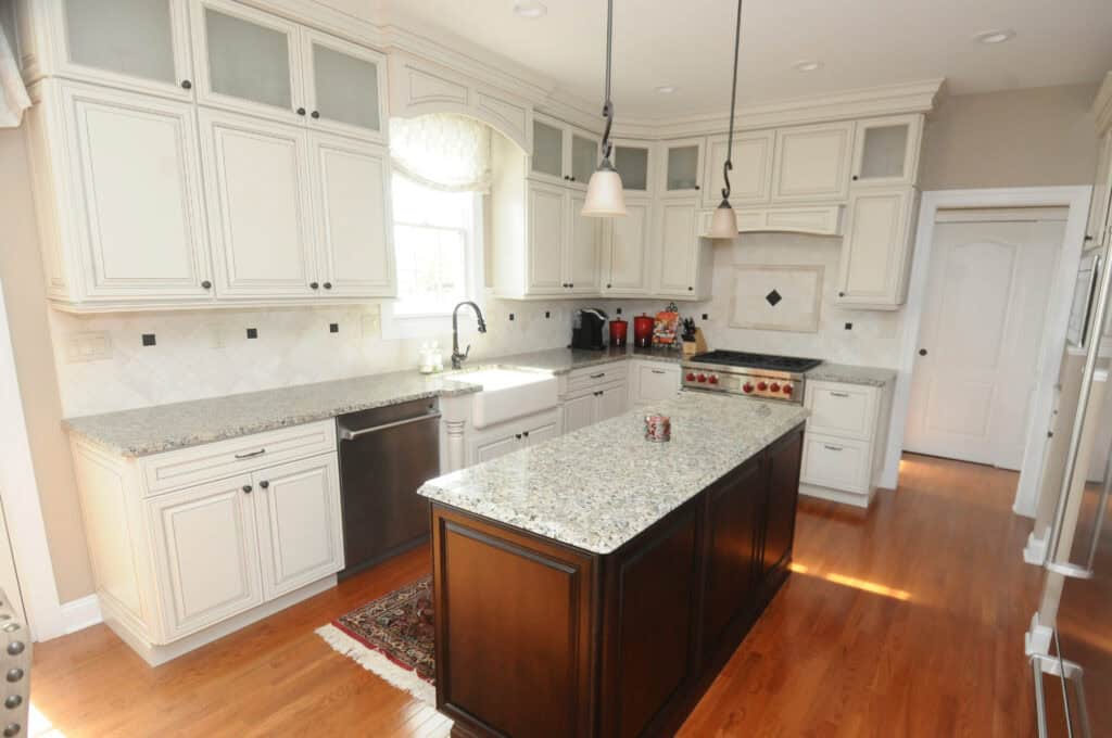 Bright kitchen with white cabinets, stainless steel appliances, granite countertops, a dark wood island, pendant lights, and hardwood floors. Sunlight streams in from a window above the sink.