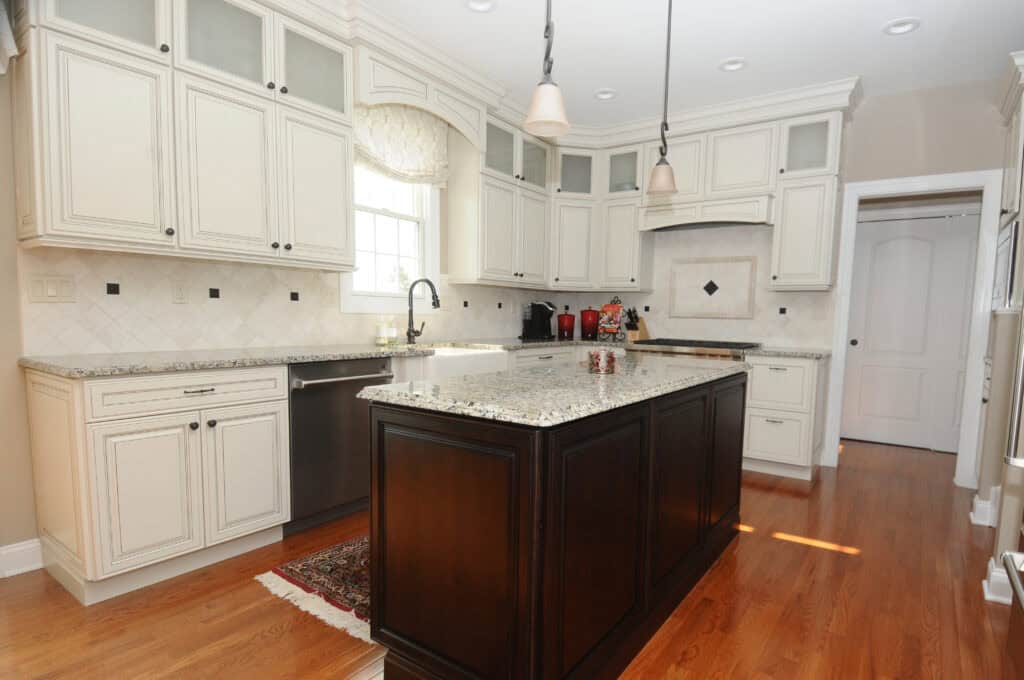 A modern kitchen with white cabinets, granite countertops, stainless steel appliances, a dark wood island, pendant lights, and hardwood floors. Sunlight shines through a window above the sink.
