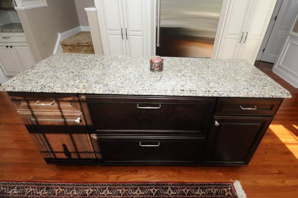 A kitchen island with a speckled granite countertop, dark wood cabinets, silver handles, and a candle in the center. The island is on a wooden floor, and rugs and stairs are partially visible in the background.