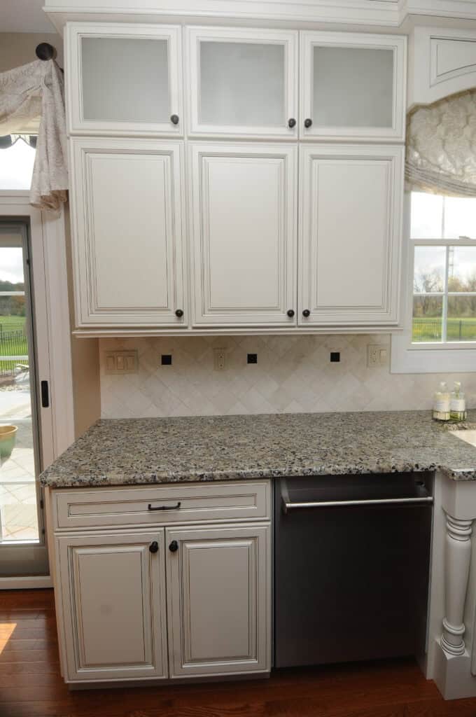 White kitchen cabinets with frosted glass panels are mounted above a granite countertop and a stainless steel dishwasher. The backsplash has a diamond tile pattern, and there is a window to the right and a door to the left.
