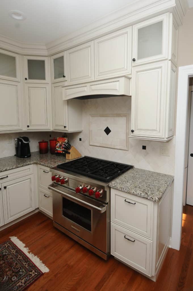A modern kitchen with white cabinets, granite countertops, a stainless steel gas stove with red knobs, and hardwood flooring. A kettle, utensil holder, and rug are also visible.