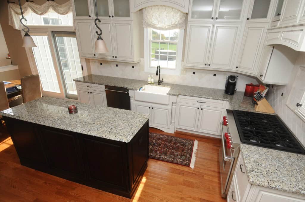 Spacious kitchen with white cabinets, granite countertops, a farmhouse sink under a window, dark island, stainless appliances, a stove with red knobs, hardwood floor, and a small rug in front of the sink.