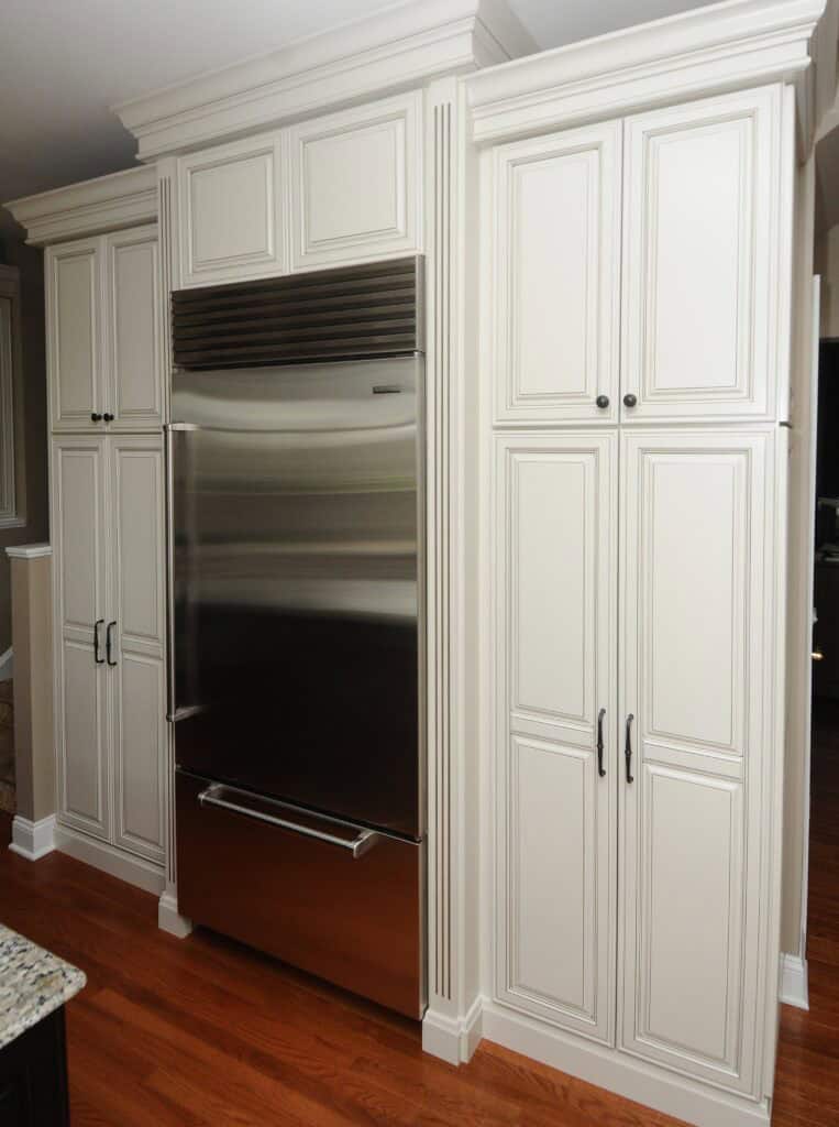 A stainless steel refrigerator is built into white paneled kitchen cabinetry with black handles, set against a background of wood floors and granite countertop.