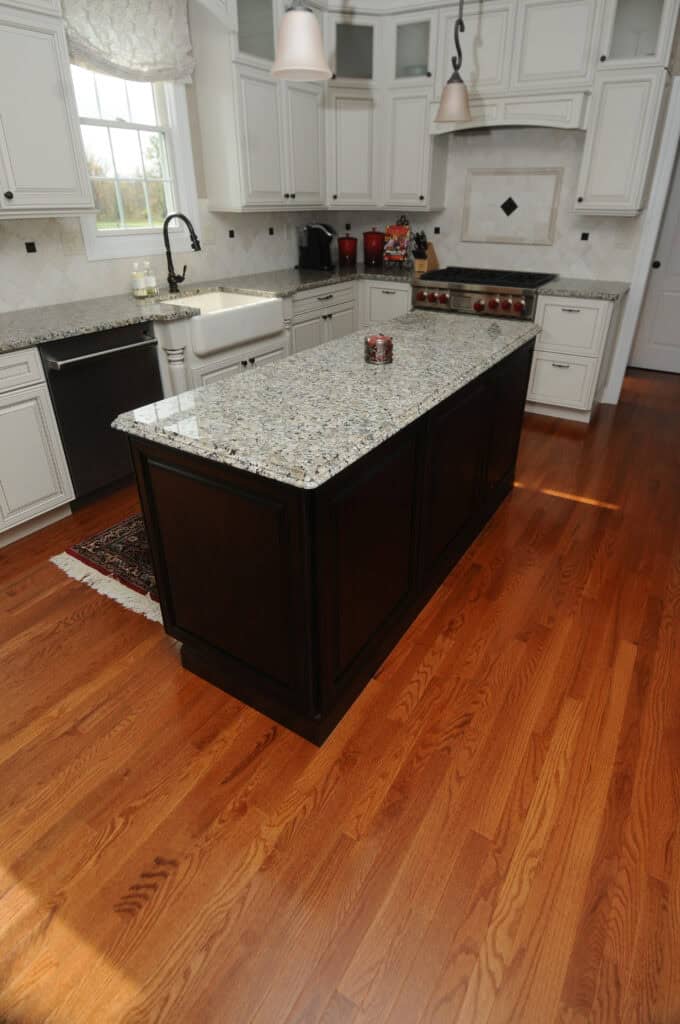 A modern kitchen with white cabinets, stainless steel appliances, a dark wood island with a granite countertop, and hardwood floors. Sunlight streams through a window above a farmhouse sink.