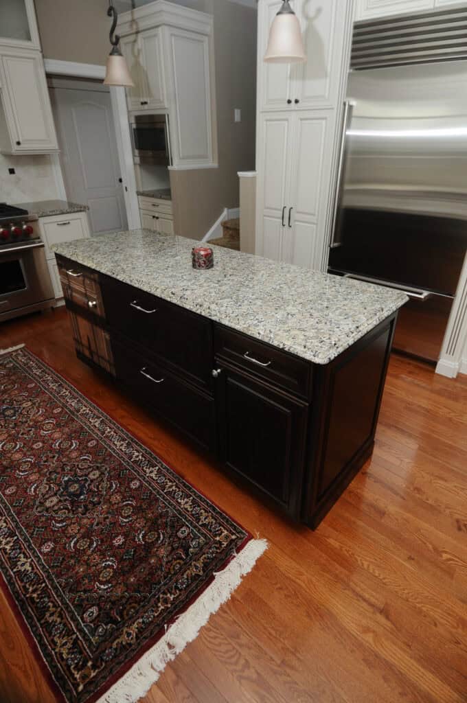 A modern kitchen with light wood floors, a patterned rug, dark cabinets, a granite island countertop, stainless steel appliances, and white cabinetry. A small decorative item sits on the island.