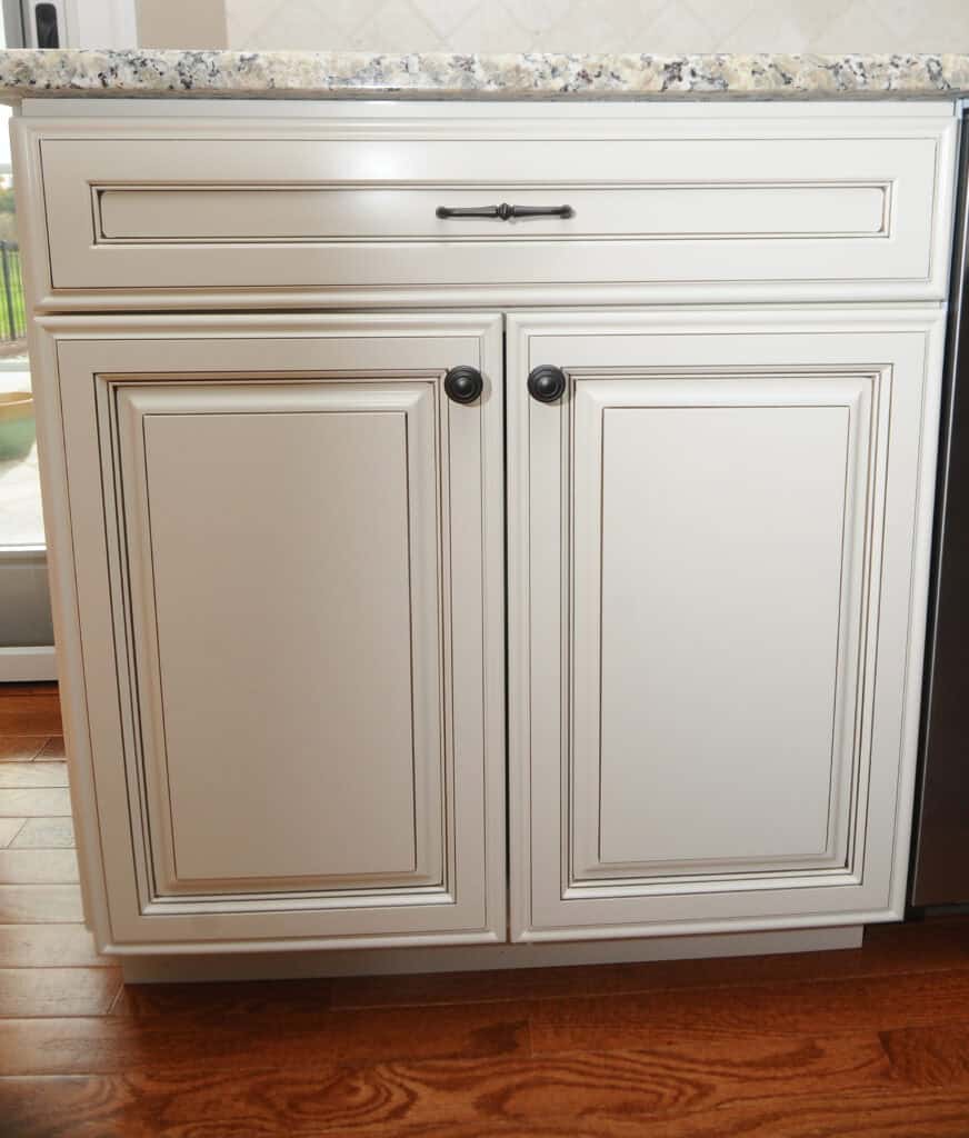 White kitchen cabinets with two doors and a drawer, featuring dark round handles and a dark drawer pull, set against a wood floor and topped with a speckled granite countertop.