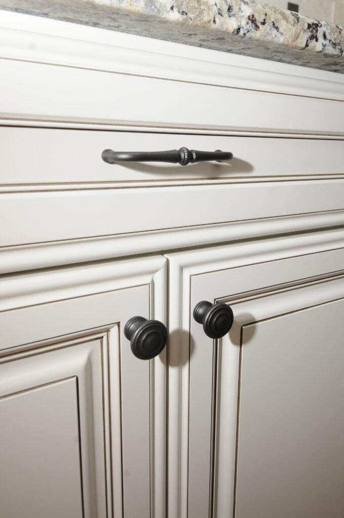 Close-up of cream-colored kitchen cabinets with black round knobs on the lower cabinet doors and a matching black handle on the top drawer, topped with a speckled granite countertop.