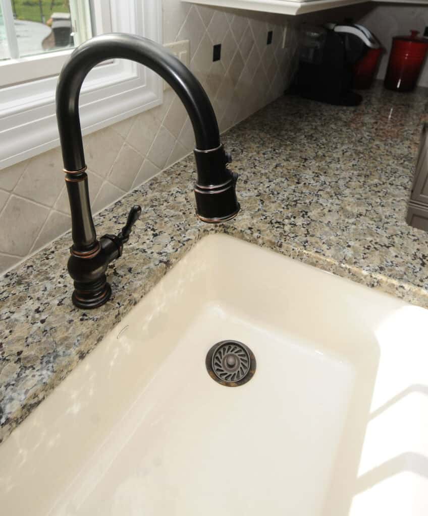 A close-up view of a kitchen sink with a black faucet, set in a granite countertop. The sink is empty, and sunlight streams in from a nearby window, casting light onto the counter and sink.