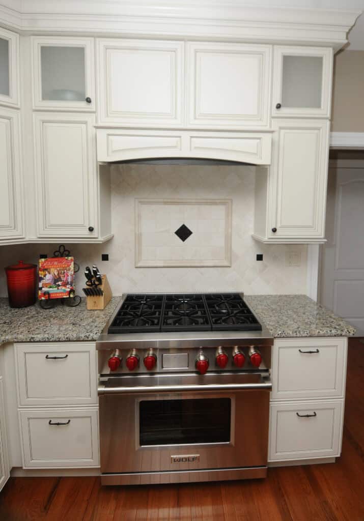 A modern kitchen with white cabinets, granite countertops, a stainless steel gas stove with red knobs, a knife block, a red canister, and a cookbook set against a tiled backsplash.