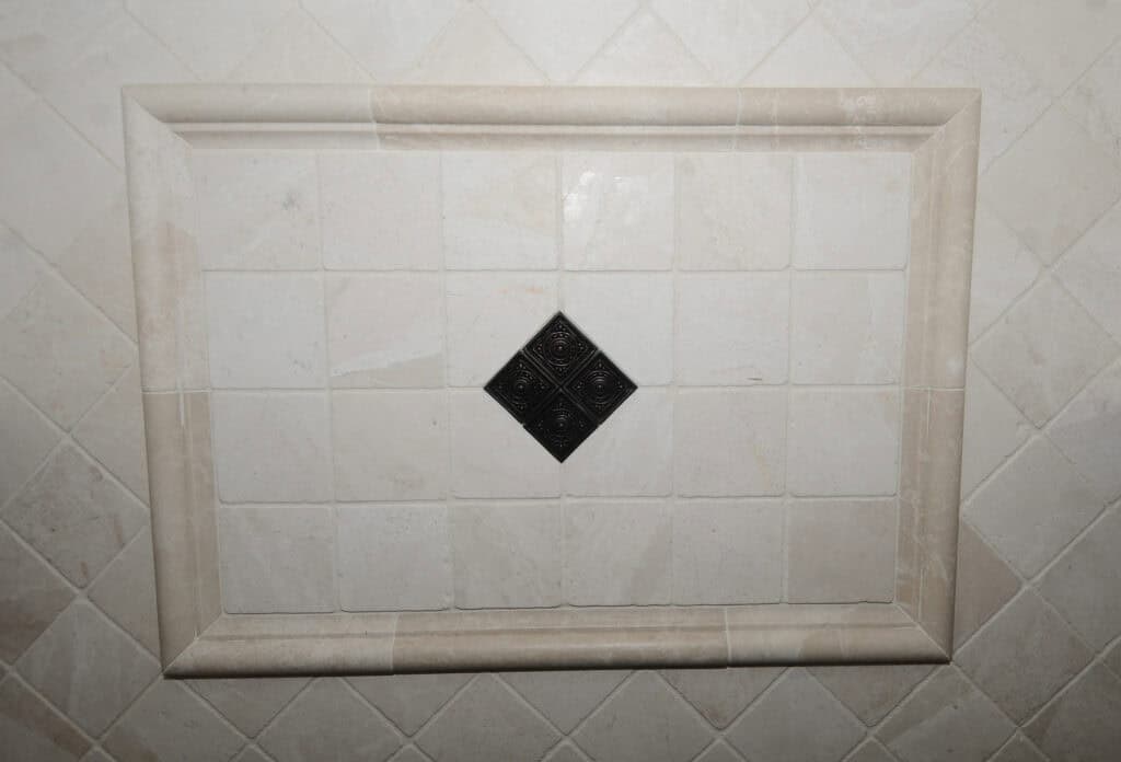 A rectangular section of beige tiled wall with a raised border and a central black diamond-shaped tile, surrounded by lighter tiles arranged in a grid pattern.