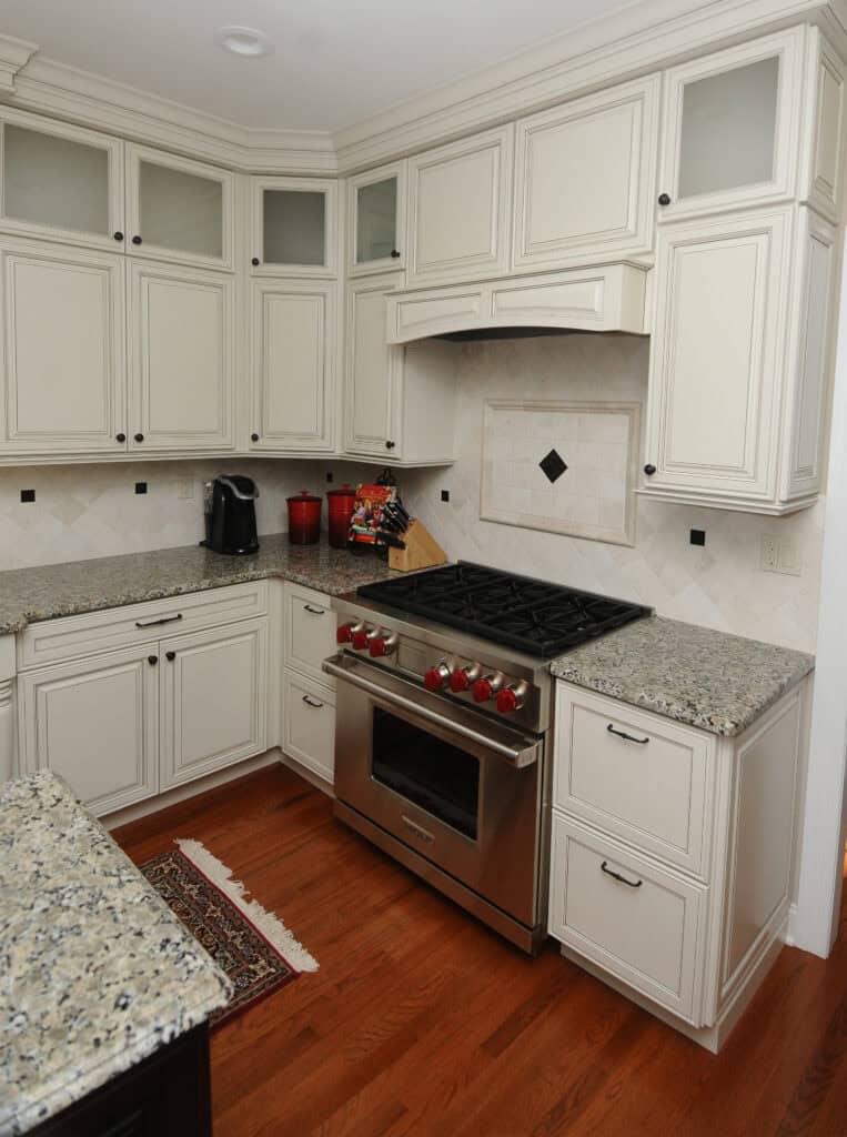 A modern kitchen with cream-colored cabinets, granite countertops, a stainless steel gas stove with red knobs, a small coffee maker, and wooden floors. A small rug lies in front of the oven.