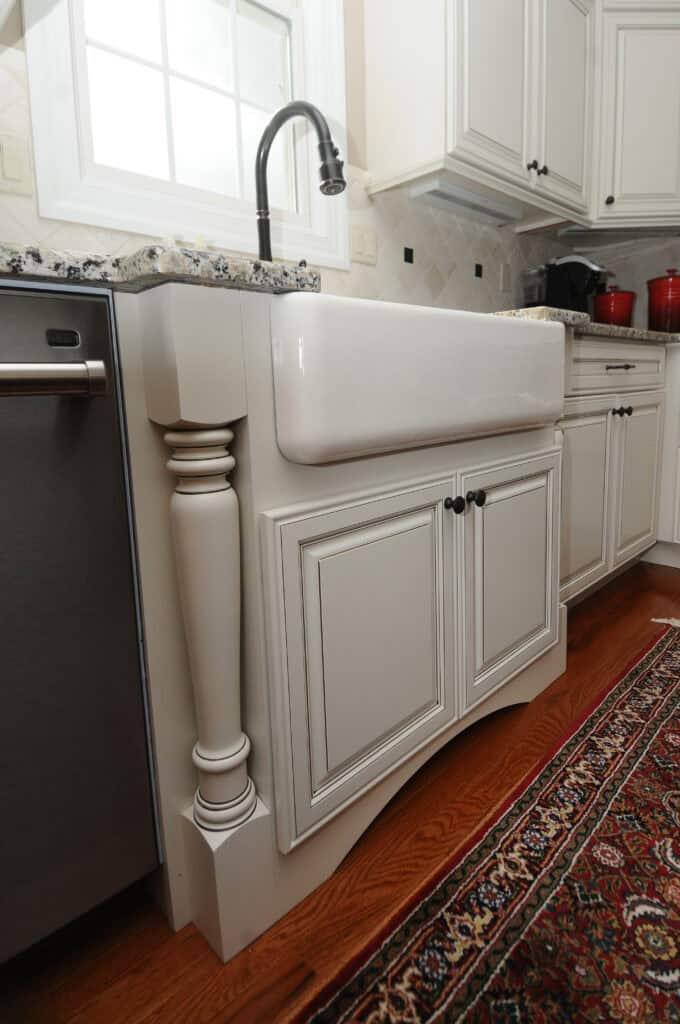 A farmhouse-style white kitchen sink with decorative cabinetry, a black faucet, and granite countertop, set against white cabinets and a dishwasher, with a patterned rug on the wooden floor.