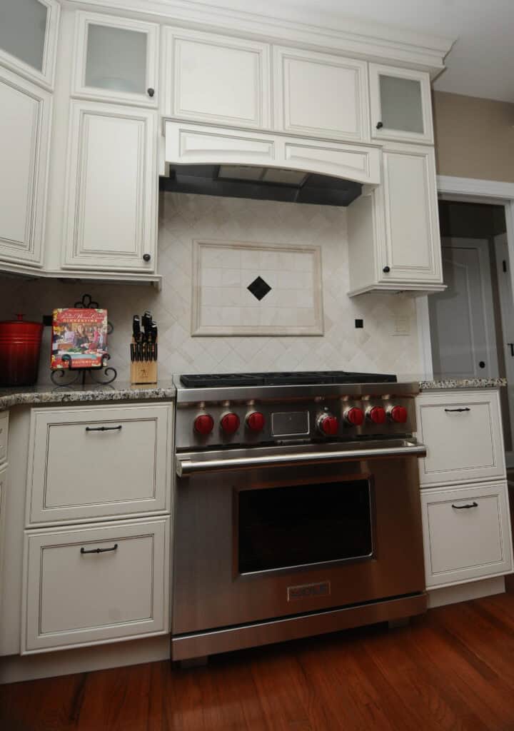 A modern kitchen features white cabinets, a stainless steel oven with red knobs, a tile backsplash with a diamond pattern, and a wooden knife block on the counter.