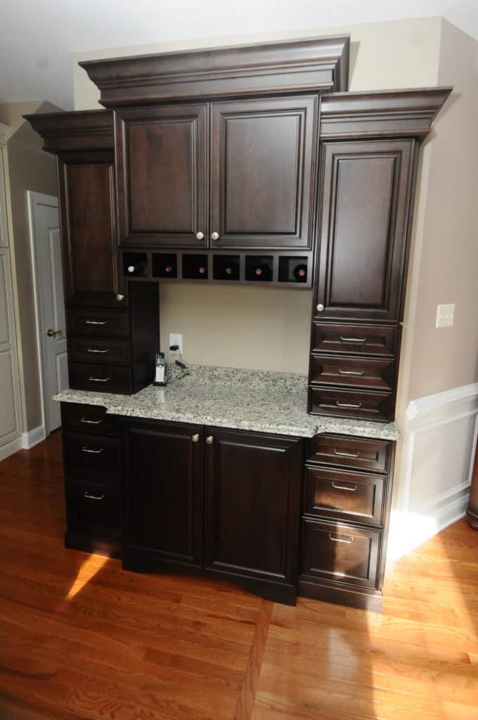 Dark wood kitchen cabinet with multiple drawers and cupboards, a granite countertop, and a built-in wine rack above the counter, set against a beige wall with wood flooring.
