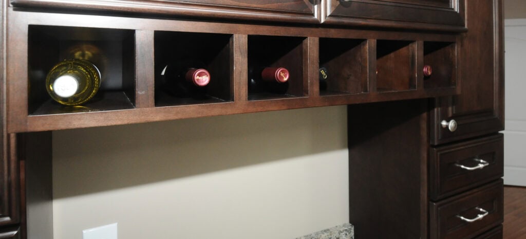 A dark wood wine rack with five cubbyholes, each holding a bottle of wine, built into kitchen cabinetry above a granite countertop.