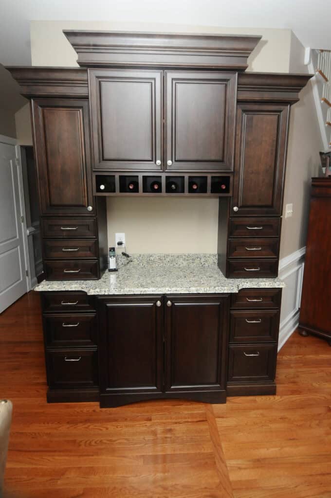 A built-in kitchen cabinet with dark wood doors and drawers, a granite countertop, and a wine rack holding six bottles. The floor is hardwood, and the background wall is light beige.