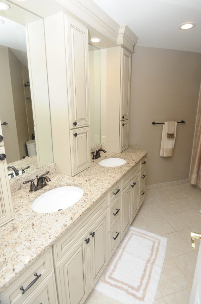 A beige bathroom features a double-sink vanity with granite countertop, cream cabinets, black fixtures, a large mirror, tiled floor, and white towels on a wall rack.