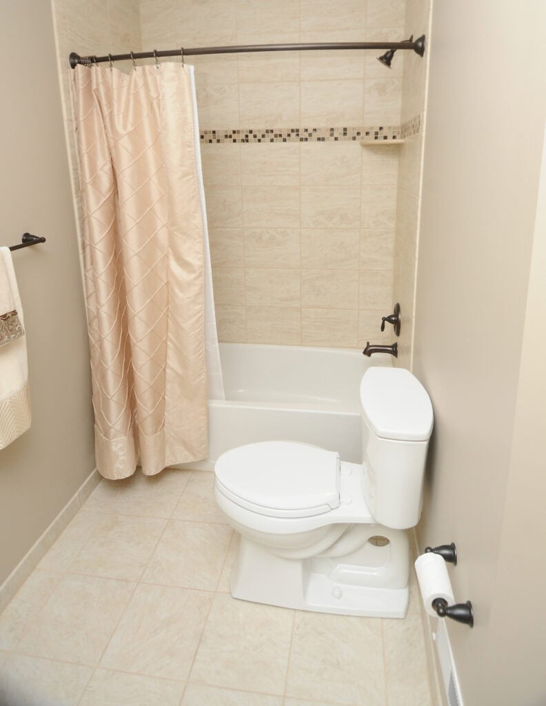 A small bathroom with beige tile, a white toilet, a bathtub with a beige shower curtain, black fixtures, and a towel and toilet paper on wall-mounted holders.