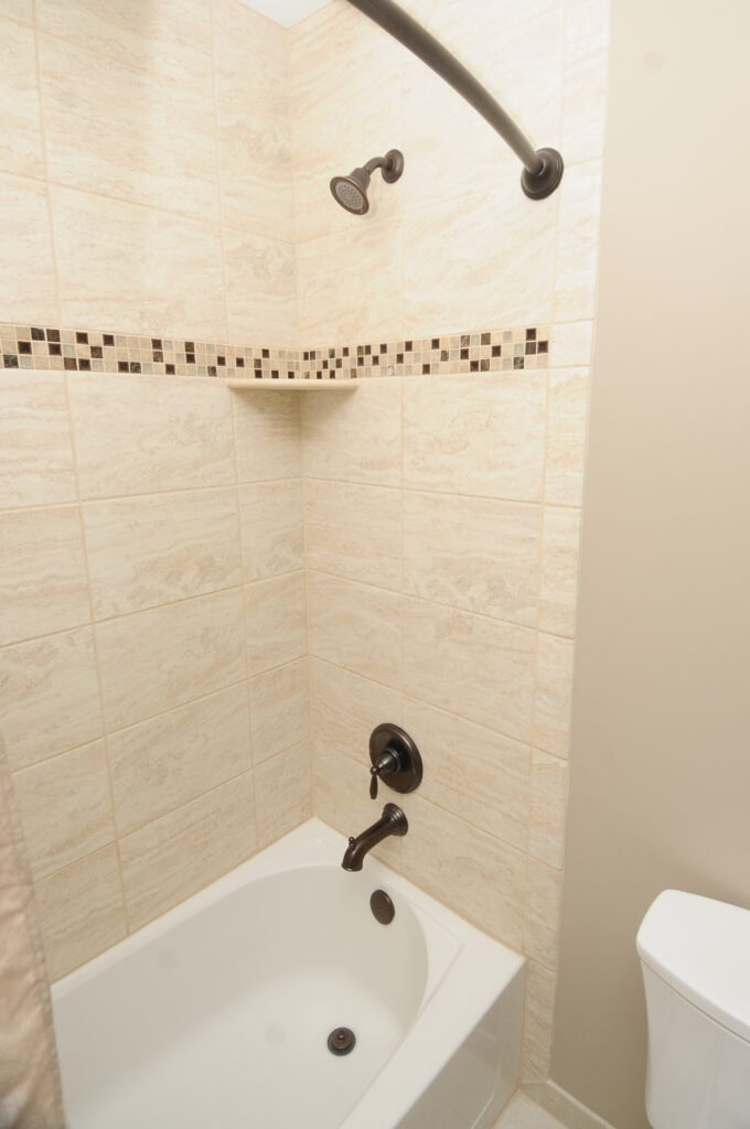 A bathroom with a white bathtub, beige tiled walls featuring a decorative mosaic border, a bronze showerhead, faucet, and handles, and a partial view of a white toilet.