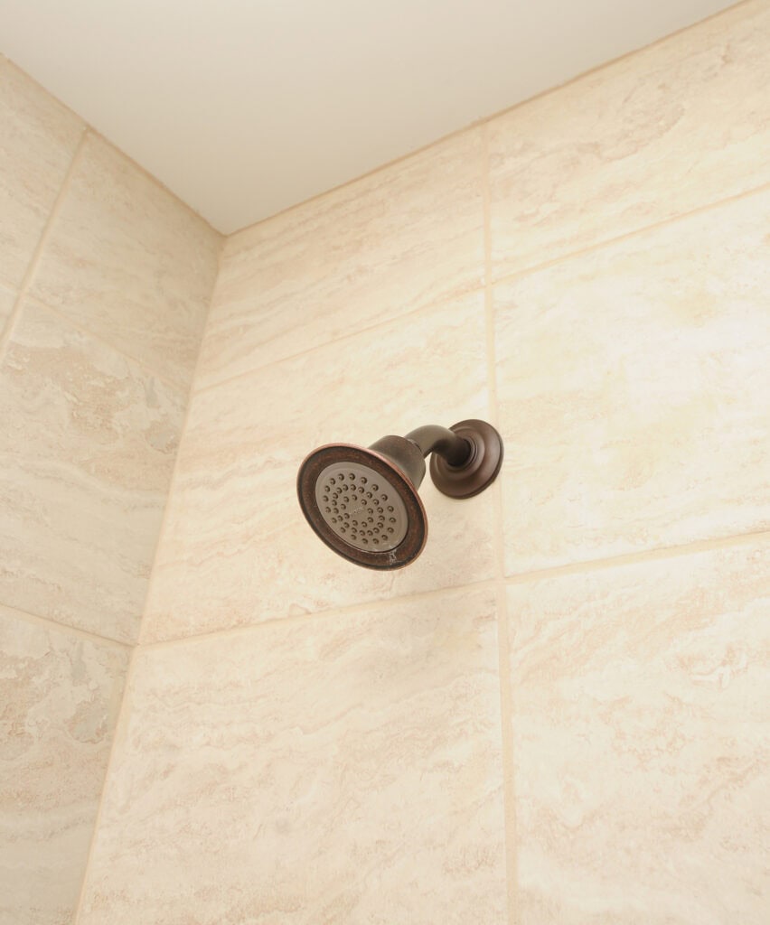 A bronze showerhead is mounted on a tiled shower wall with a beige and cream marble pattern. The photo is taken from below, showing the showerhead and part of the ceiling.