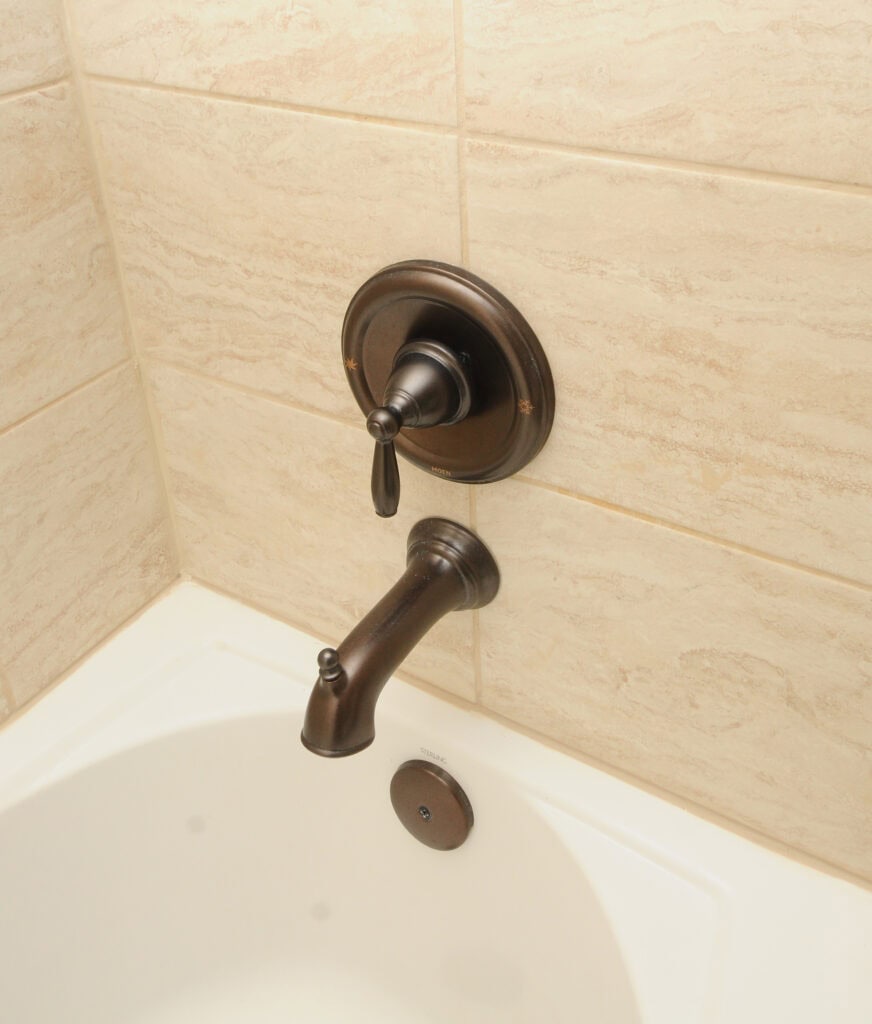 Bronze bathtub faucet with matching single-handle control mounted on a beige tiled wall above a white bathtub. The tub also has a circular drain cover visible near the faucet.