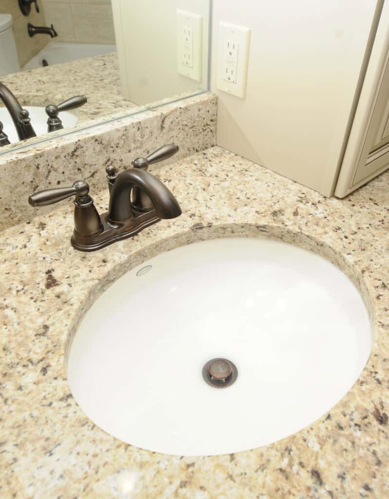 Oval white bathroom sink with a dark bronze faucet set in a speckled beige granite countertop. A mirror, electrical outlets, and part of a cabinet are also visible in the background.