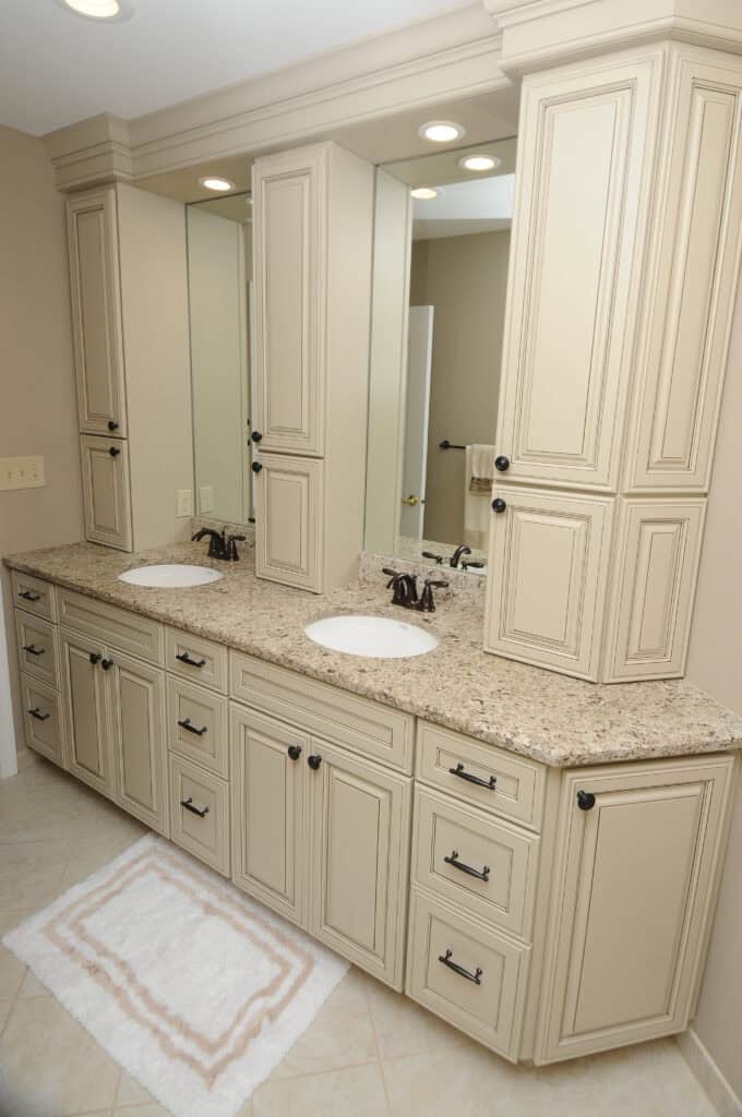 Cream-colored bathroom vanity with granite countertop, dual sinks, bronze faucets, and large mirrors; surrounded by tall cabinets and drawers. A white textured rug lies on the tiled floor in front of the vanity.