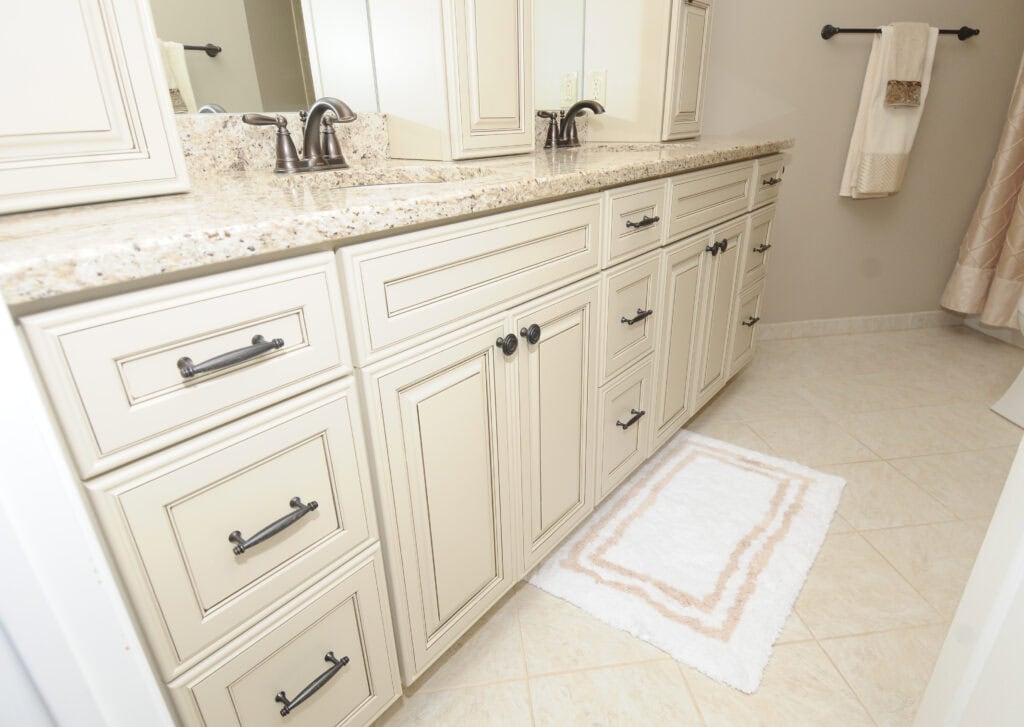 A bathroom with cream-colored cabinets, granite countertop, two sinks with dark faucets, a white and beige rug on tiled floor, and a towel hanging on the wall.