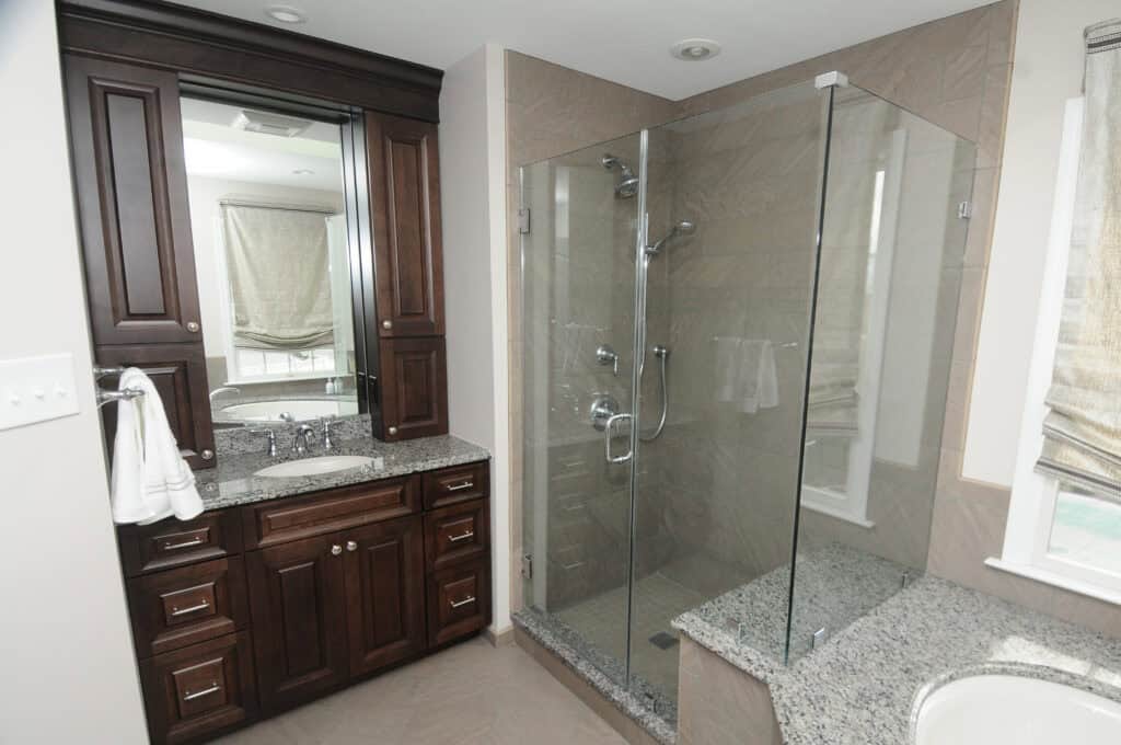 Modern bathroom with dark wood vanity, granite countertop, built-in sink, large mirrored cabinet, and a glass-enclosed shower with chrome fixtures. Beige tile floor and walls, window with light-colored shade.