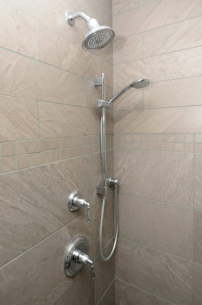 A modern shower with beige tile walls, featuring a rainfall showerhead, a handheld showerhead on a sliding bar, and two chrome control handles.