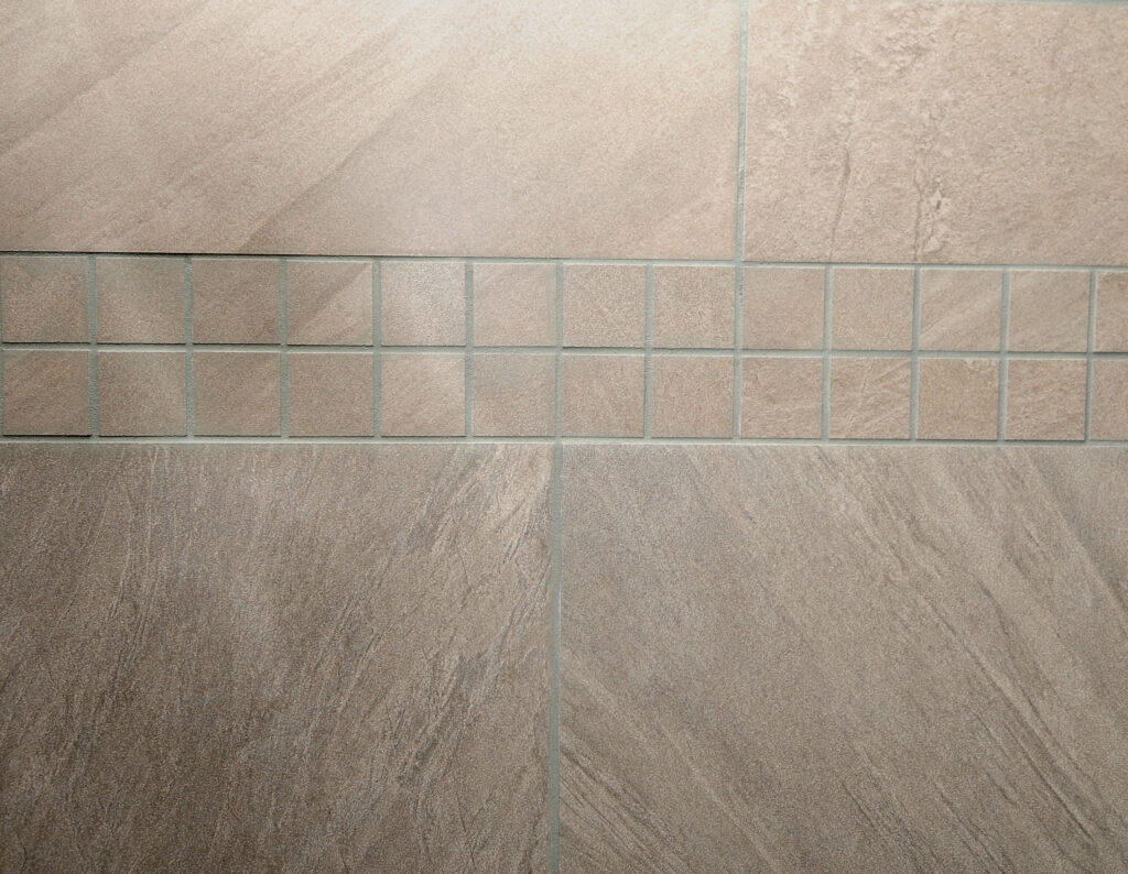 Close-up of a tiled wall with large rectangular beige tiles, bordered by a row of small square tiles in the center, all separated by light-colored grout lines.