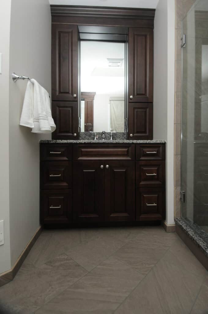 A modern bathroom with dark wood cabinets, a granite countertop, a large mirror, white towels on a silver towel rack, and a glass shower on the right. The floor has large beige tiles.