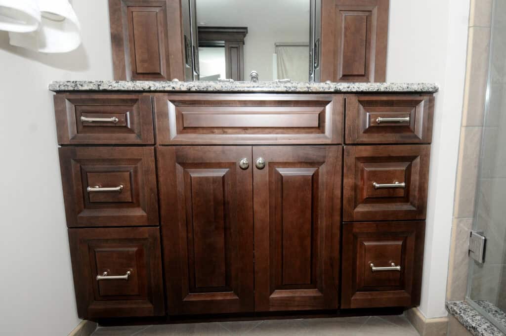 A wooden bathroom vanity with a granite countertop, silver handles on drawers and cabinets, and a sink partially visible with a mirror above. The vanity is set against a light-colored wall.