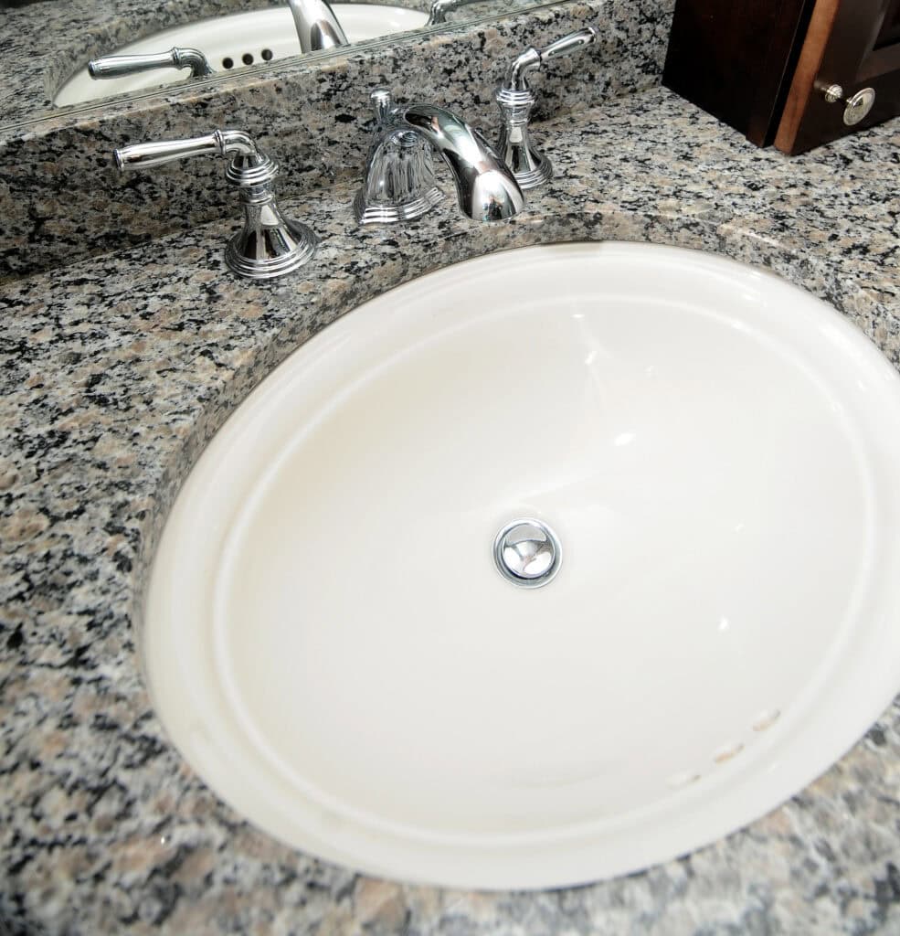Oval white sink with a silver drain and chrome faucet set in a speckled granite countertop, with a matching granite backsplash and a partial view of a wooden item on the right side.