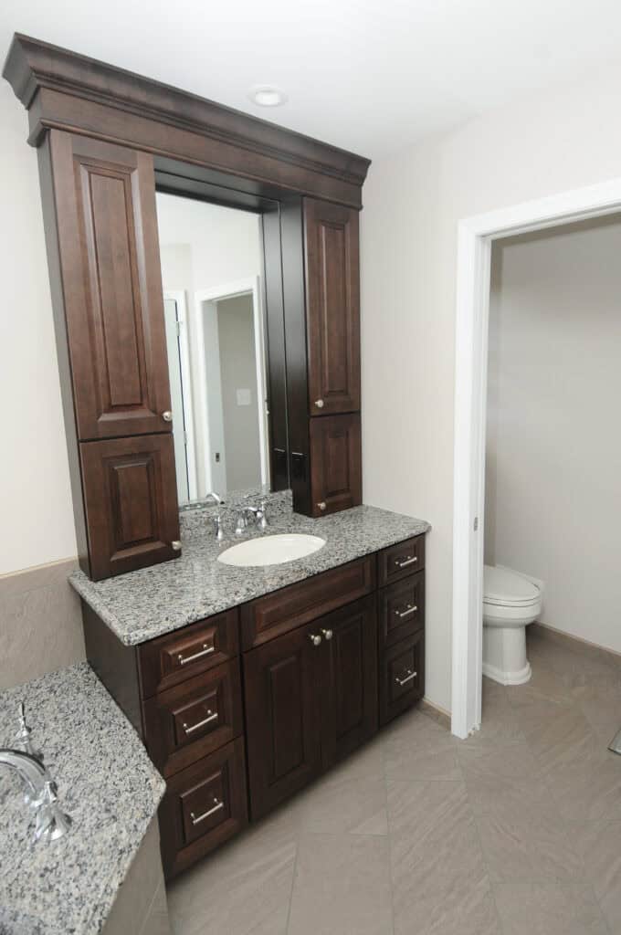 A bathroom with dark wood cabinets, granite countertops, an undermount sink with a chrome faucet, a large mirror, and a separate toilet area visible through an open doorway.