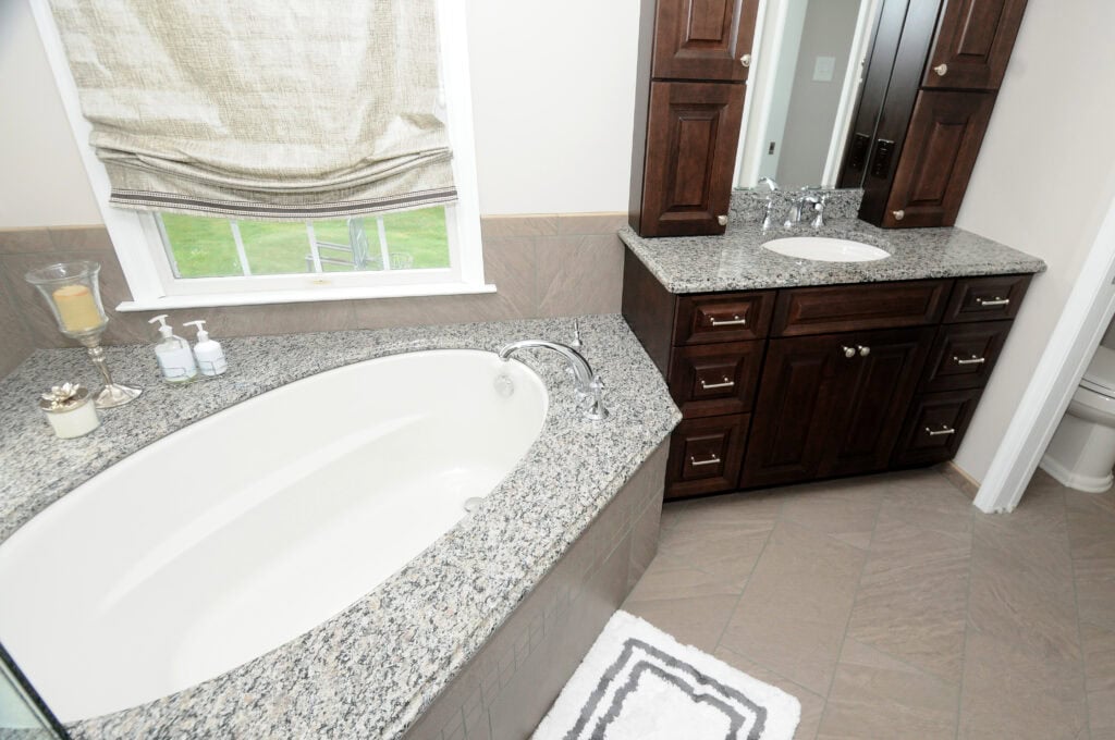 A bathroom features a large oval bathtub with granite surround, a granite-topped vanity with a sink, dark wooden cabinets, a window with a fabric shade, tile flooring, and a white bath mat.