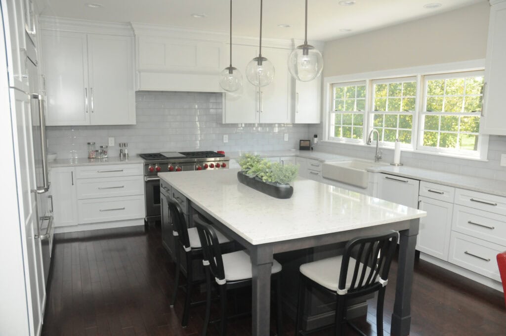 Modern kitchen with white cabinets, gray subway tile backsplash, large island with white countertop, black chairs, stainless steel appliances, farmhouse sink, pendant lights, and windows letting in natural light.