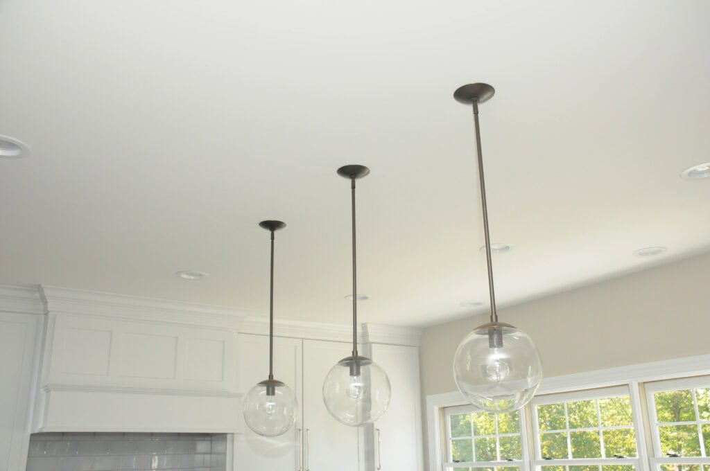 Three modern pendant lights with clear glass globes hang from a white ceiling in a bright room with large windows and white cabinetry. Sunlight streams in through the windows.