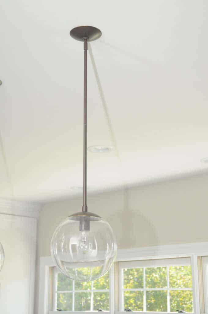 A clear glass globe pendant light hangs from the ceiling by a long, thin rod. Sunlight streams through nearby windows, illuminating the light fixture and casting soft shadows.